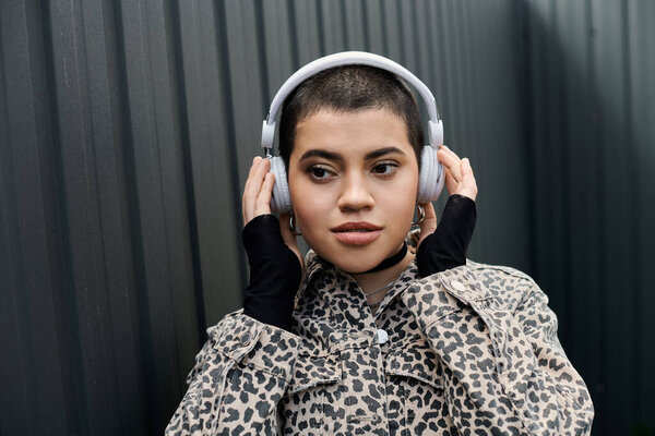 Стильная молодая женщина с короткими волосами в наушниках и леопардовой куртке рядом с современным зданием.