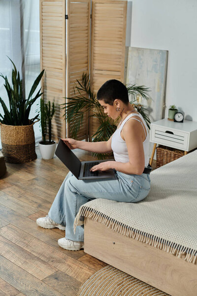 Молодая женщина с короткими волосами в повседневной одежде, сидящая на кровати, погруженная в использование ноутбука.