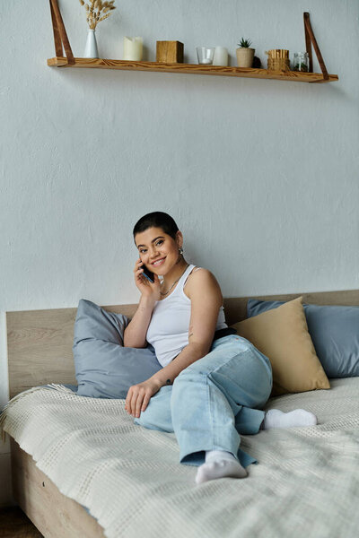 Молодая женщина с короткими волосами в повседневной одежде сидит на кровати, разговаривая по мобильному телефону.