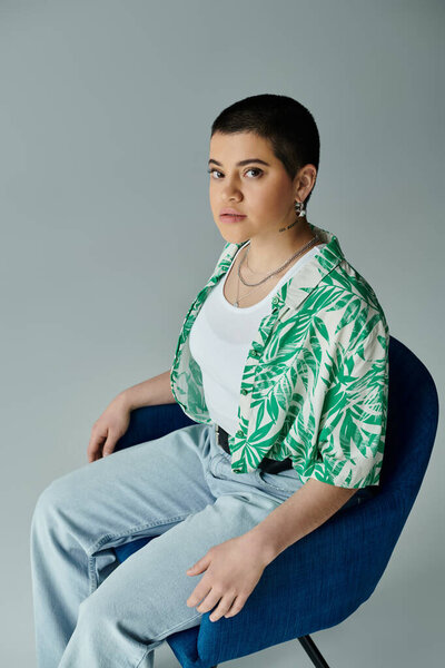 Стильная молодая женщина с короткими волосами расслабляется на синем стуле, одетая в шикарную зеленую и белую полосатую рубашку.