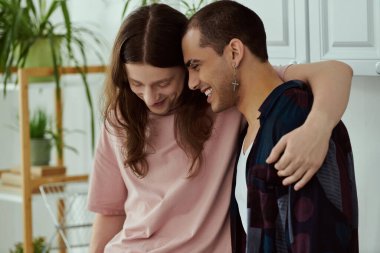 Eşcinsel bir çift, gelişigüzel giyinmiş, yan yana durmuş, birbirlerine şefkat ve sevgiyle sarılıyorlar..