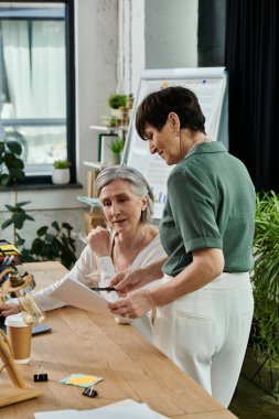 Two women, one standing, one sitting, working together at a table in an office setting. clipart