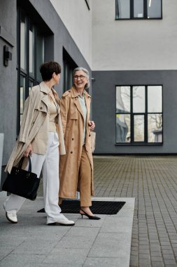 Modern bir binanın önünde iş kıyafeti giymiş iki yetişkin kadın..