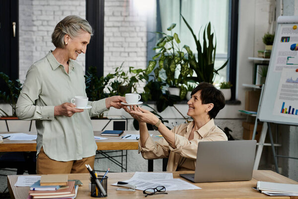 A woman passes a cup of coffee to another woman in an office setting.
