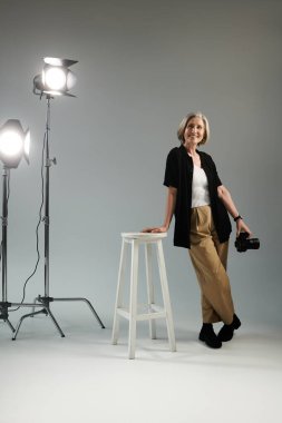 Orta yaşlı bir kadın, parlak bir stüdyo ışığının önündeki taburede kamerayla poz veriyor.