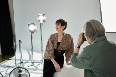 A middle-aged lesbian couple in a photo studio; one taking a picture with a camera while the other poses as a model. clipart