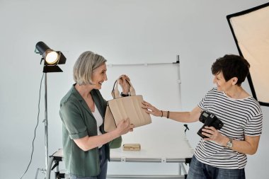 Profesyonel bir fotoğraf stüdyosunda orta yaşlı bir lezbiyen çift, birinin elinde çanta, diğerinin elinde kamera var..