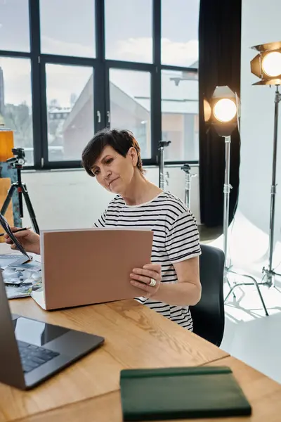 stock image A middle-aged woman sits at a desk in a modern studio, working on a laptop.