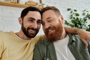 Two men, both with beards, relax on a couch and hugging at home clipart