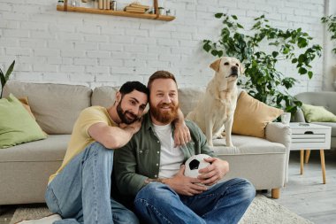 Sakallı iki adam kanepeye oturup spor maçı seyrediyor. Arka planda Labrador köpeği var..