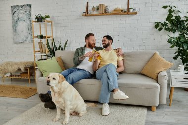 İki adam, sakallı eşcinsel bir çift, bir kanepede oturup labrador köpeğiyle oturma odasındaki spor maçını izliyorlar..