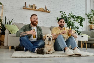 Sakallı iki adam kanepede oturup Labrador köpeğiyle oturma odasında maç izliyor..