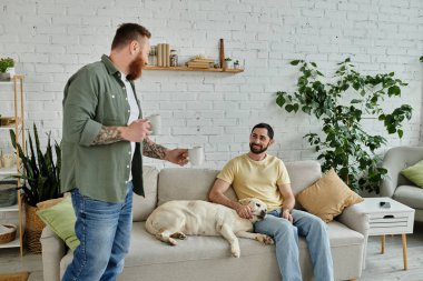 İki adam, biri bardaklı, biri kanepede oturuyor, oturma odasında labrador köpeğiyle birlikte..