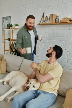 Sakallı bir adam, rahat bir oturma odasında labrador köpeğinin yanında, erkek arkadaşının elinde fincanlarla rahat bir şekilde oturur.