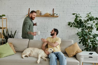 Sakallı adam ve labrador köpeği rahat bir oturma odasında kanepede rahatlar, erkek arkadaşı elinde fincanlarla