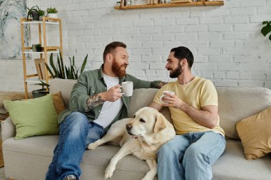 Sakallı bir çift, sıcak bir oturma odasında, bir labrador köpeğinin yanında bir kanepede oturup kahve içiyorlar.