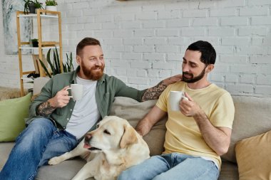 Sakallı eşcinsel bir çift, sıcak bir oturma odasında Labrador köpeği eşliğinde bir kanepede oturuyor..