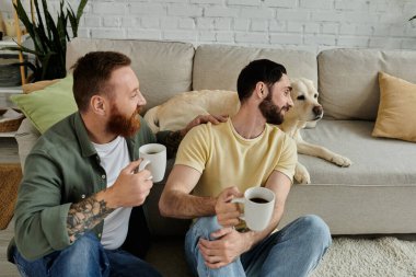 İki adam, sakallı eşcinsel bir çift, oturma odalarında bir Labrador köpeğinin yanında kahve kupaları tutuyorlar..