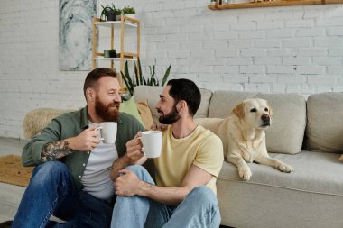 İki adam, sakallı eşcinsel bir çift, rahat bir oturma odasında labrador köpeğinin yanında bir kanepede oturuyorlar..