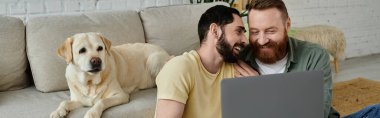 Bearded gay couple and their Labrador dog sitting on a couch, both engrossed in viewing content on a laptop. clipart