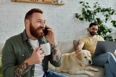 A bearded man sits on a couch, talking on a cell phone, working remotely beside his partner and a labrador dog in a cozy living room. clipart