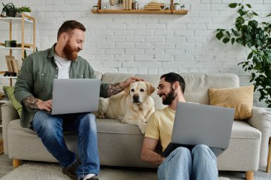 İki sakallı adam rahat bir oturma odasında Labrador köpeğiyle kanepede otururken dizüstü bilgisayarlarda çalışıyor..