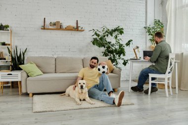 Sakallı eşcinsel adam, rahat oturma odasında yanında iki labrador köpeği otururken uzaktan çalışıyor..
