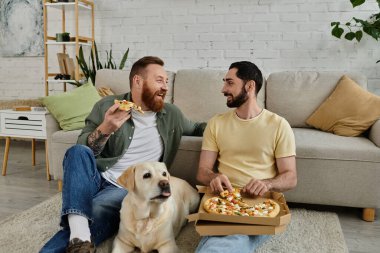 Sakallı eşcinsel çift, Labrador köpekleri ile birlikte oturma odasında pizzanın tadını çıkarıyor..