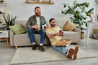 İki sakallı adam kanepede rahatlar, sıcak oturma odasında pizza ve bira paylaşırlar..