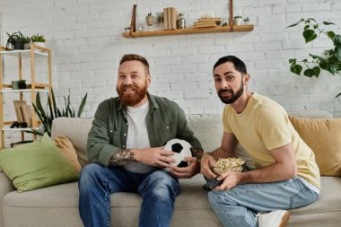 İki adam, sakallı eşcinsel bir çift, kanepede oturup rahat bir oturma odasında futbol topu tutuyorlar..