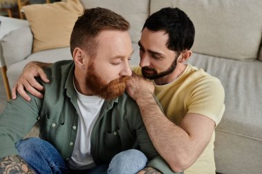 A cozy scene of a gay couple with beards, sitting closely together on the backrest of a couch in their living room. clipart