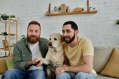 Sakallı eşcinsel çift, tüylü arkadaşları Labrador 'la evde rahat bir kanepede uzanarak eğleniyor..