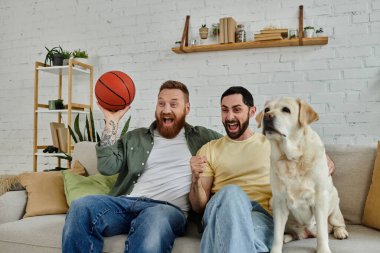 İki sakallı adam rahat bir oturma odasında, dost canlısı bir Labrador köpeğinin yanındaki koltukta dinleniyor..
