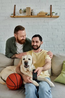 Bearded gay couple enjoying a cozy moment on a couch with their labrador in a living room. clipart