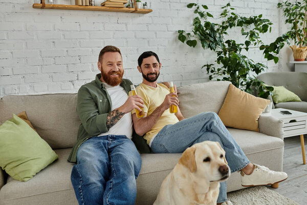 Двое мужчин, гей-пара, сидят на диване со своей собакой и вместе смотрят спортивный матч в своей гостиной.