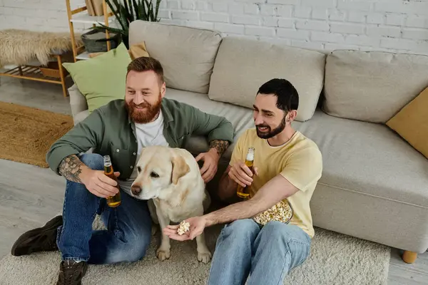 Sakallı eşcinsel çift, rahat oturma odasında Labrador köpeğiyle spor maçı seyrediyor..