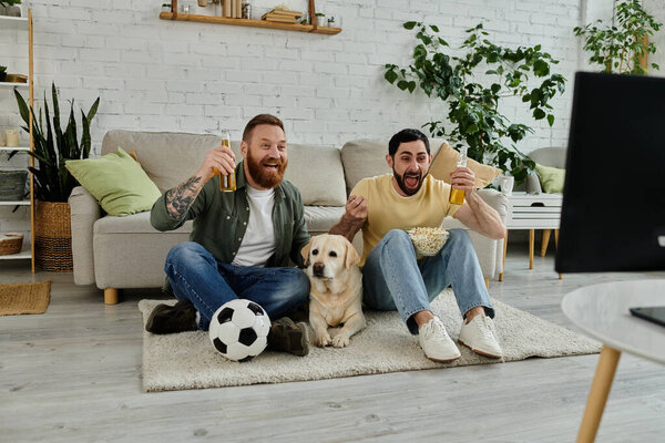 Двое мужчин с бородой сидят на ковре с собакой-лабрадором и смотрят спортивный матч в гостиной..