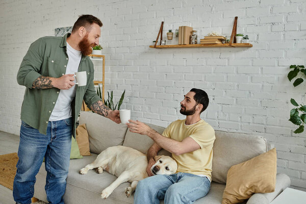 Bearded man sitting on a couch next to a labrador dog, his partner offering drink