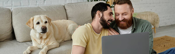 Bearded gay couple and their Labrador dog sitting on a couch, both engrossed in viewing content on a laptop.