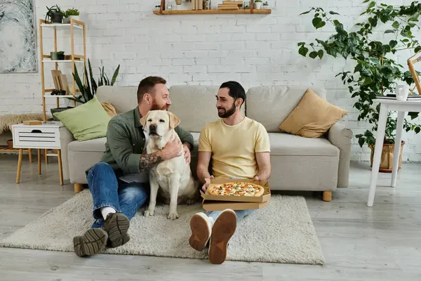 stock image Two men with beards relax on a couch with a box of pizza, sharing a slice while a friendly Labrador looks on.