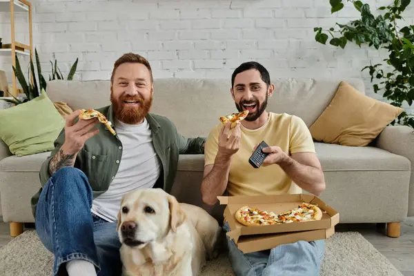 Stock image Two bearded men enjoy pizza on a couch with their labrador dog in the living room.