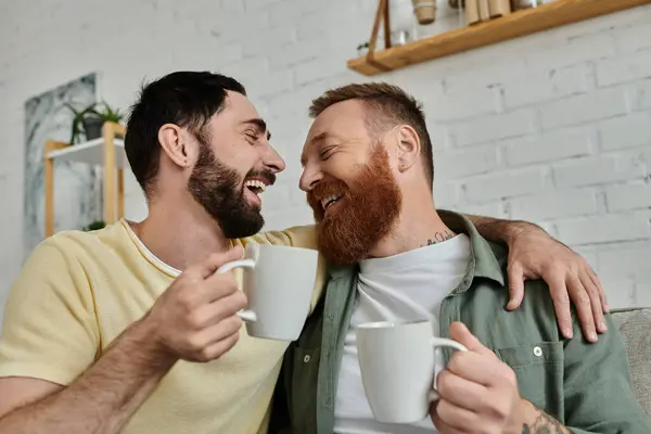 stock image A bearded gay couple sits closely on a couch, enjoying each others company in their cozy living room.