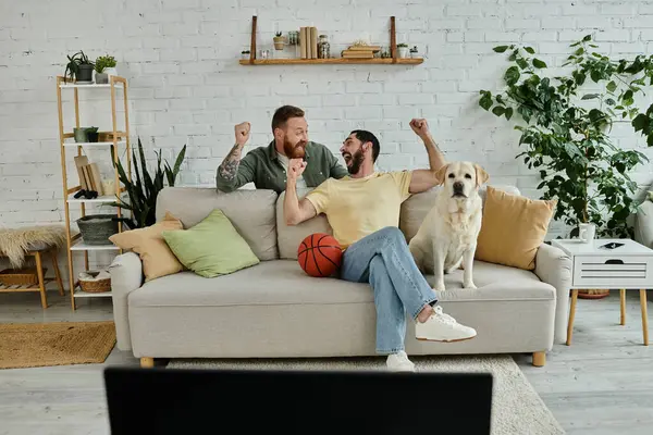 stock image Two men, one with a beard, sitting on a couch with a dog, enjoying a relaxing moment in their living room.