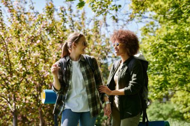 A young lesbian couple hikes through a forest on a sunny spring day. They are both wearing backpacks and smiling at each other, enjoying the beautiful scenery. clipart