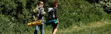 Two young women, one with long blonde hair and the other with short afro hair, hike through a wooded trail, carrying backpacks and rolled-up sleeping mats. clipart