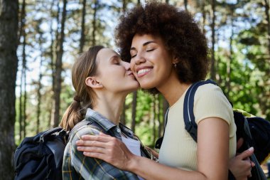 Two young women, one with dark curly hair and one with long blonde hair, share a loving embrace in a wooded area. clipart
