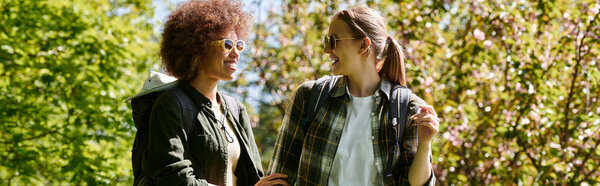 Two young women, dressed in casual outdoor wear, hike through a forest on a sunny day.