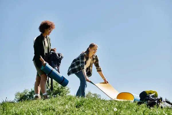 stock image Two young women prepare for a camping trip in the wilderness, rolling out a sleeping pad on the grassy ground.