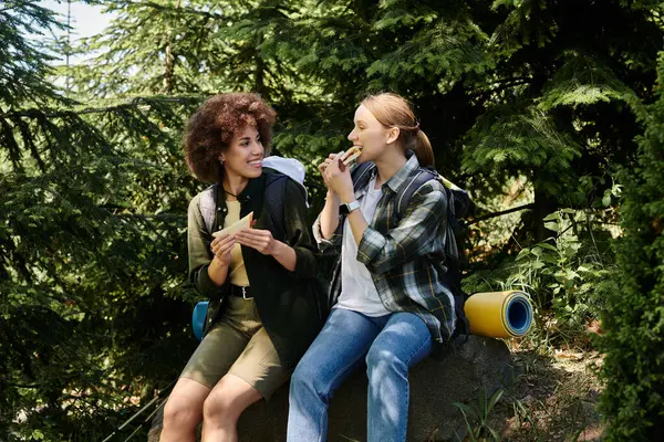 stock image Two young women, a lesbian couple, enjoy a snack break while hiking in a lush green forest.