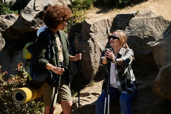 stock image Two young women, one with a backpack and trekking poles, hike together through a rocky terrain.